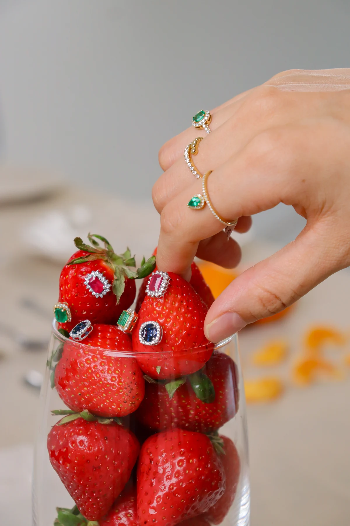 Anillos con piedras de colores sobre fresas y una mano con joyería.